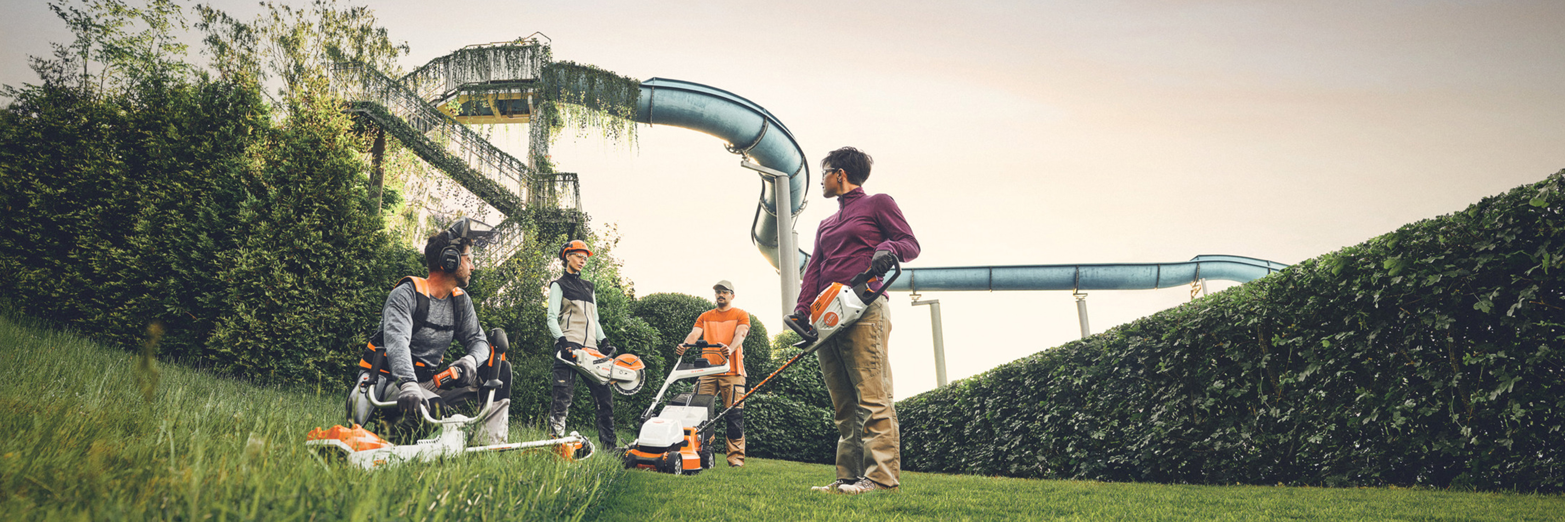Ryhmä ihmisiä työskentelee STIHL-puutarhatyökaluilla vihreällä alueella, jossa on suuri pensasaita ja vesiliukumäkiä taustalla. Heillä on käytössään raivaussahat, ruohonleikkurit ja suojavarusteet.