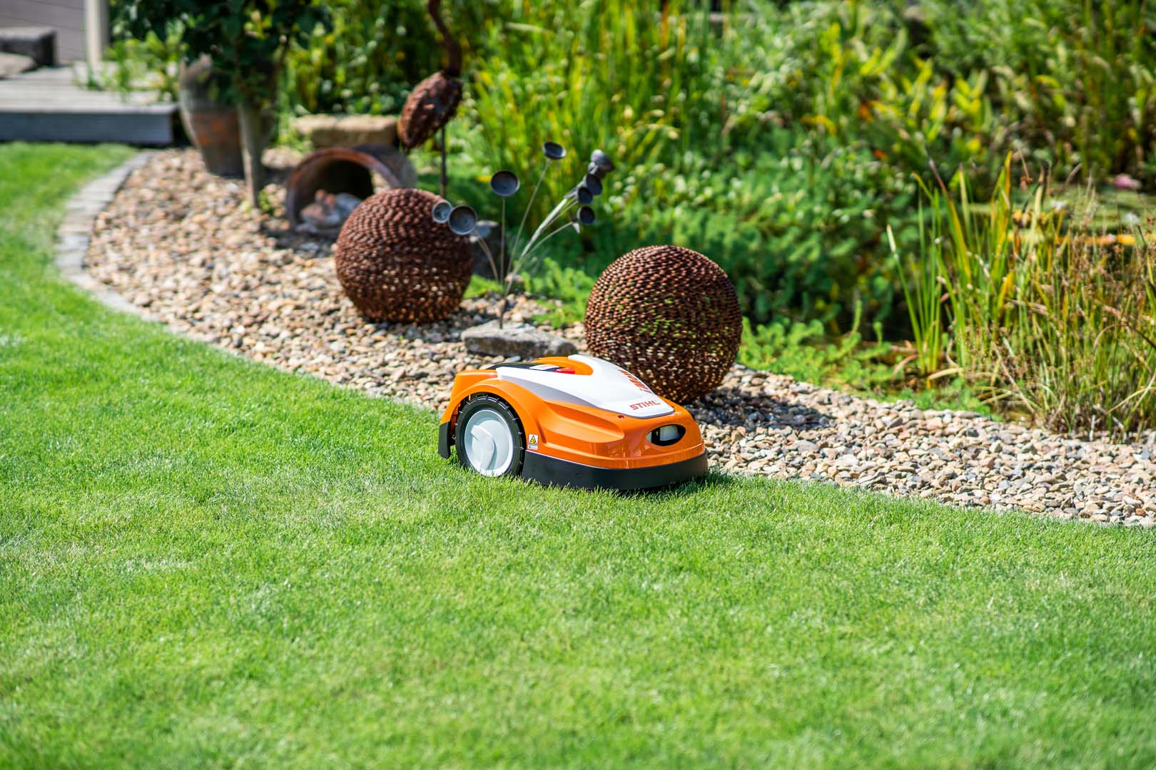 STIHL iMOW® 422 -robottiruohonleikkuri nurmikolla sorapenkin vieressä, jossa on puutarhaveistoksia
