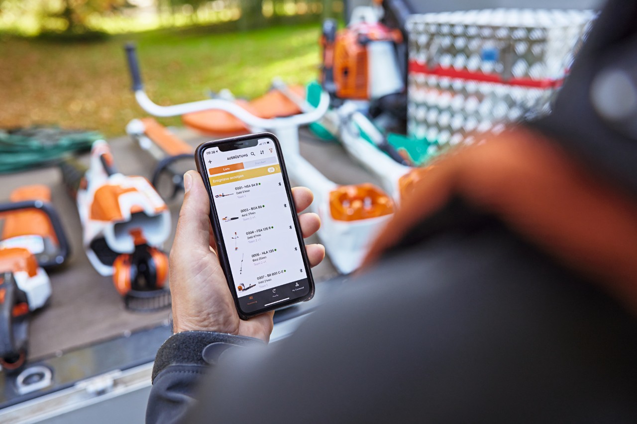 Henkilö seisomassa lava-auton lastausalueen edessä, kun alueella on erilaisia STIHL-työkaluja. Henkilö pitämässä älypuhelinta kädessään STIHL connected -sovelluksen ollessa auki. 