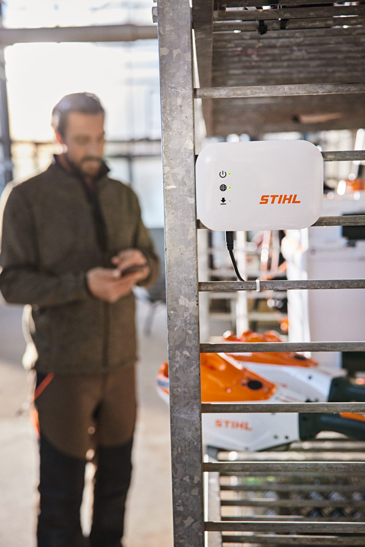 STIHL connected Box ripustettuna työpajan hyllylle. Taustalla puutarhuri ja maisemasuunnittelija vuorovaikutuksessa älypuhelimensa kanssa. 