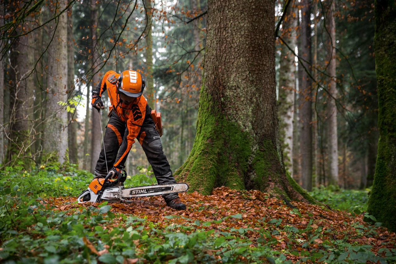 Suojavarusteisiin pukeutunut mies käynnistämässä polttomoottorikäyttöistä STIHL MS 500i -moottorisahaa lehtimetsässä