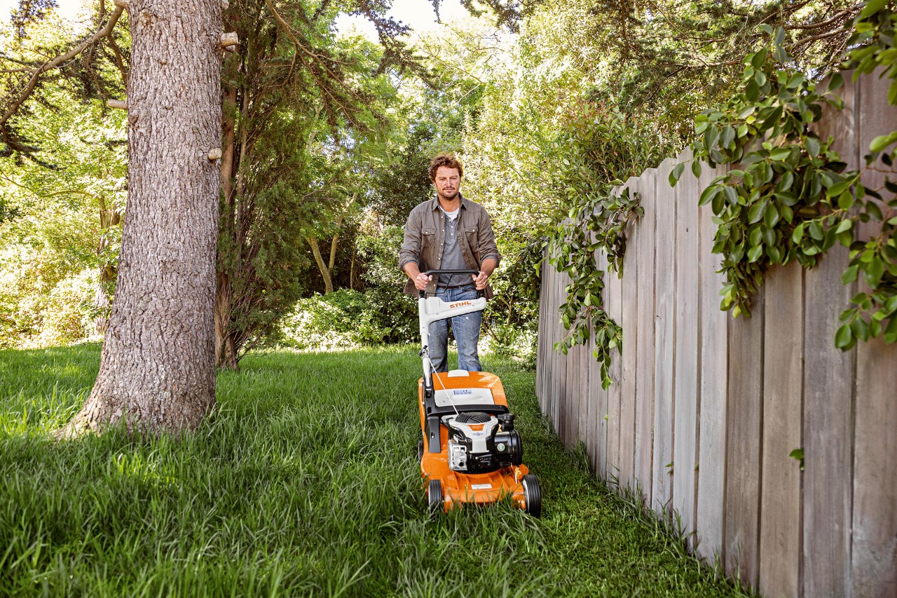 Mies leikkaa nurmikkoa polttomoottorikäyttöisellä STIHL-ruohonleikkurilla RM 448 puuaidan vieressä