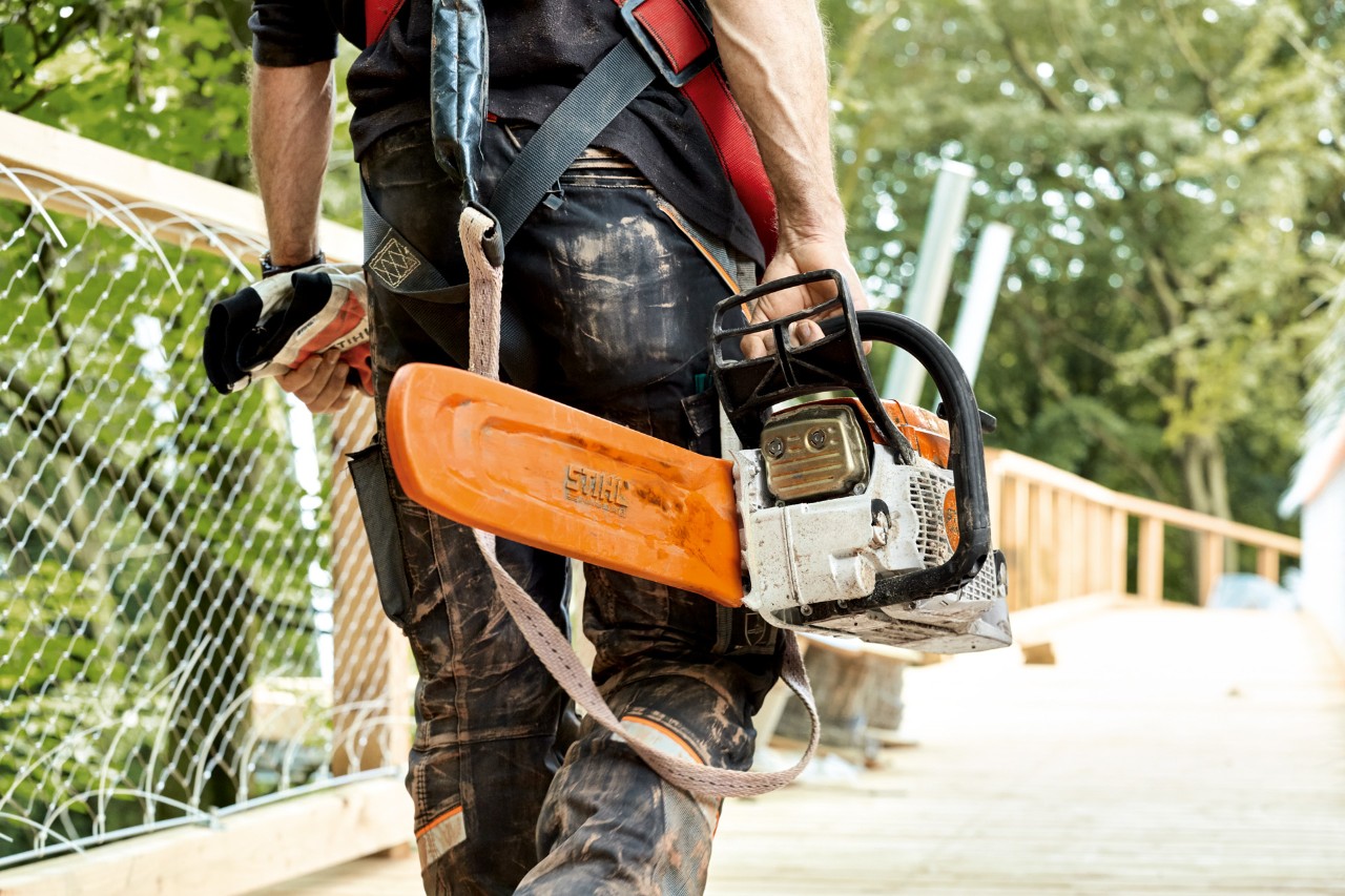 Bensiinikäyttöinen ketjusuojuksella varustettu STIHL MS 261-moottorisaha, jota mies kantaa kävellessään sillan yli