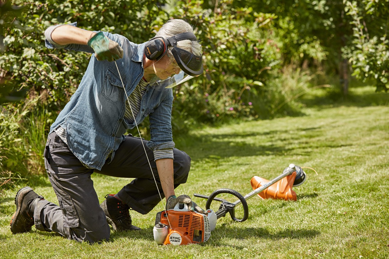 Nainen käyttämässä kuulon- ja kasvosuojaimia käynnistäessään bensiinikäyttöistä STIHL FS 56 R -raivaussahaa nurmikolla