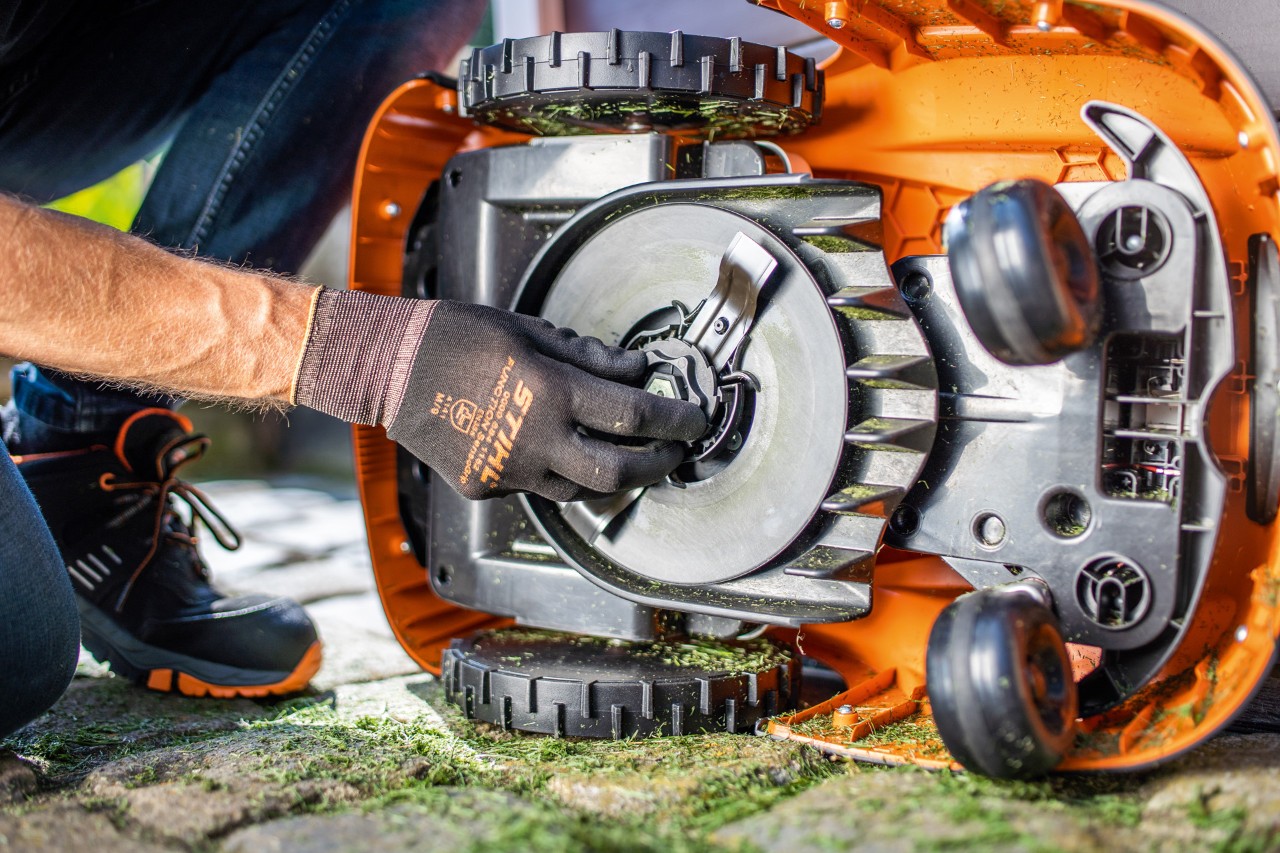 STIHL iMOW® 422 -robottiruohonleikkuri kallistuu sivulleen, kun käsineet kädessä oleva henkilö tarkastaa terää