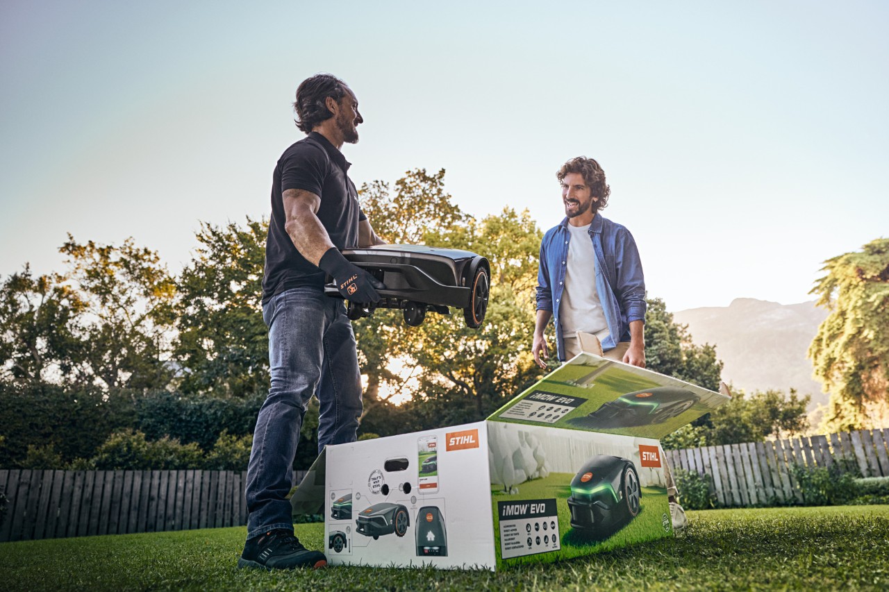 STIHL-jälleenmyyjä seisomassa asiakkaan puutarhassa iMOW® EVO kädessään ja iMOW®-laatikko edessään, valmiina auttamaan asennuksessa
