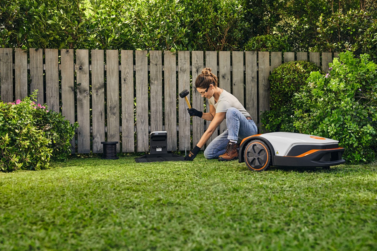 Nainen asentamassa STIHL iMOW® -leikkurin telakointiasemaa puutarhassaan aidan edessä.