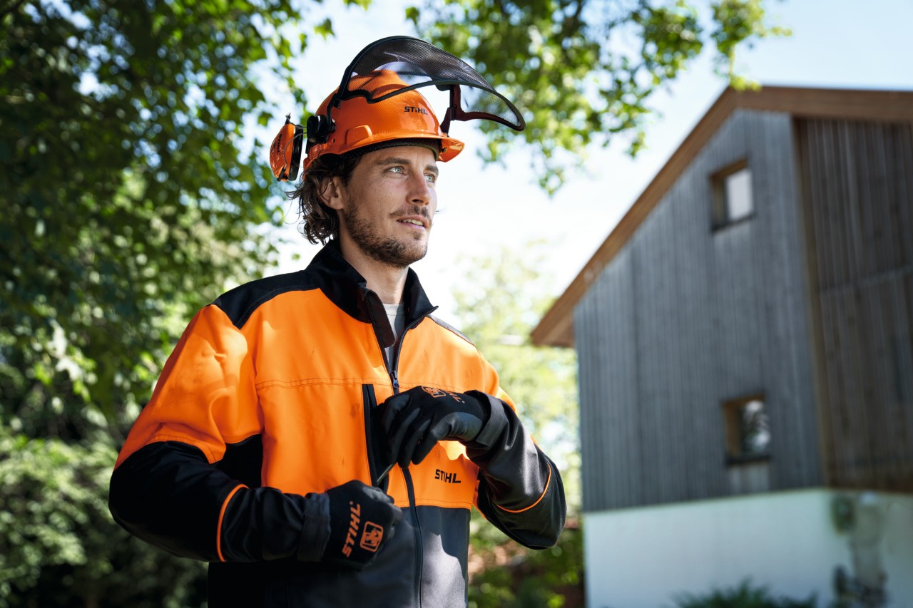 Mies, jolla on oranssit ja mustat STIHL-suojavarusteet, mukaan lukien kypärä ja käsineet, katsoo itsevarmasti kuvasta poispäin, taustalla talo ja puita 