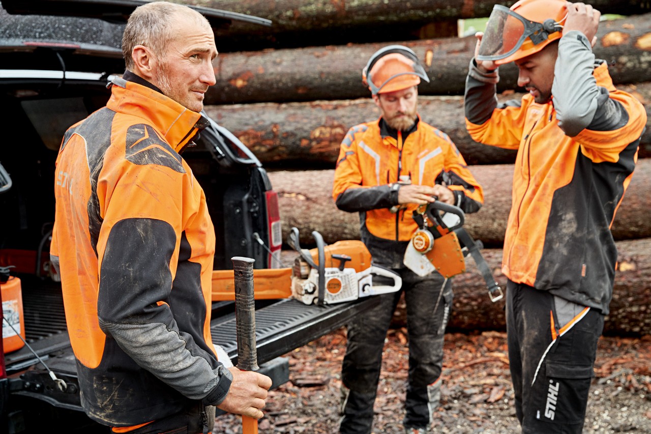 Kolme miestä STIHL-suojavaatteissa valmistautumassa käyttämään STIHL-moottorisahaa puupinon edessä