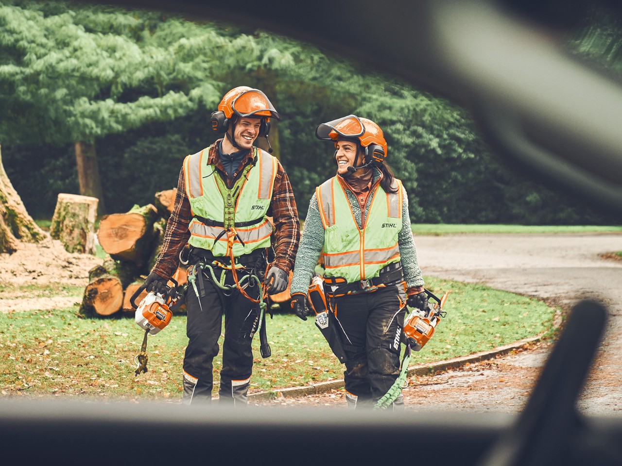 Mies ja nainen, joilla on mukanaan STIHL akkukäyttöiset moottorisahat ja STIHL akkukäyttöinen oksasakset, kävelevät hymyillen kohti autoa.  